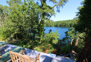 A deck with wooden chairs and a table overlooking a lake surrounded by dense trees. - Private Cottage Compound On Ryders Pond with Pristine Yard (2215) (Truro)