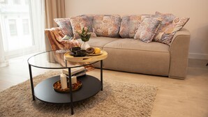 Una sala de estar con un sofá color beige, cojines estampados, una mesa de centro de cristal con una bandeja de aperitivos y un jarrón con flores.