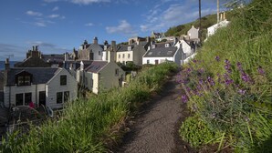 A narrow gravel path leading through a residential area with houses on both sides, surrounded by greenery and purple flowers. - Ellie-Jo Cottage (Gardenstown)