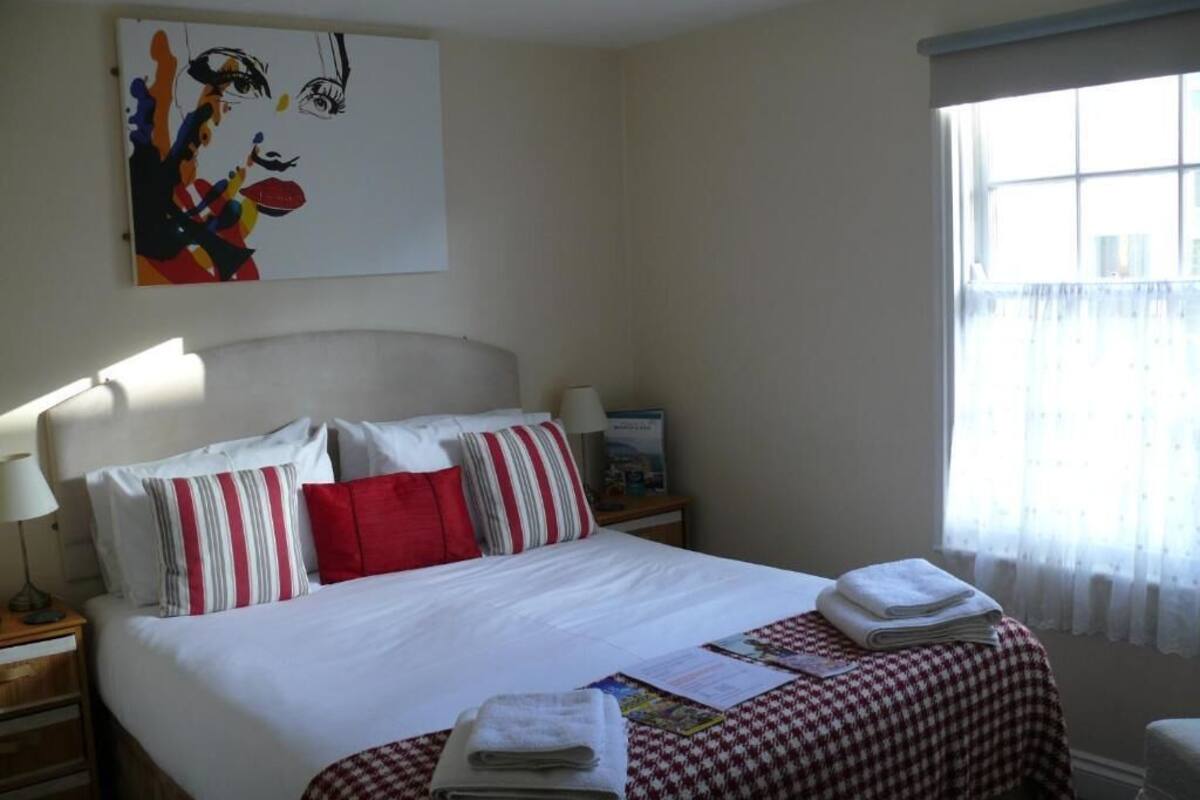 A hotel room with a large bed, striped pillows, a checkered bedspread, a window with sheer curtains, and a painting on the wall.