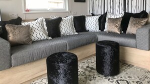 A modern living room with a sectional sofa, patterned cushions, a black ottoman, a window with a view, and a wall with a geometric pattern.