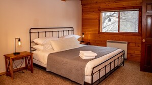 A bedroom with a wooden cabin-style interior, a metal bed frame, a bedside table with a lamp, and a window with a view of trees. - 3BR Pet Friendly | Mountain Views, Dark Skies, Walk to Trails, Exceptional Coffee (Pagosa Springs)