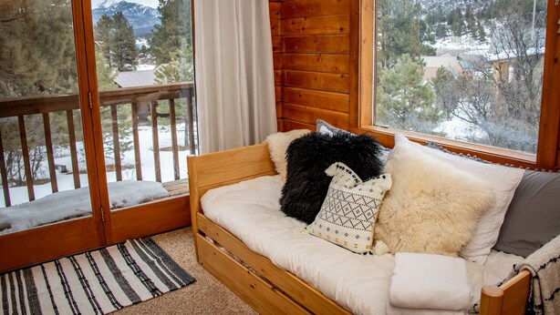 A cozy cabin room with a wooden interior, a sofa with cushions, a view of snowy mountains, and a wooden deck outside. - 3BR Pet Friendly | Mountain Views, Dark Skies, Walk to Trails, Exceptional Coffee (Pagosa Springs)