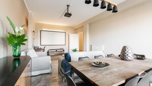 Ein modernes Wohnzimmer mit einem Holzesstisch, einer Couch und einem Couchtisch. An der Wand ist ein Projektionsschirm angebracht, und eine Deckenleuchte sorgt für Licht.