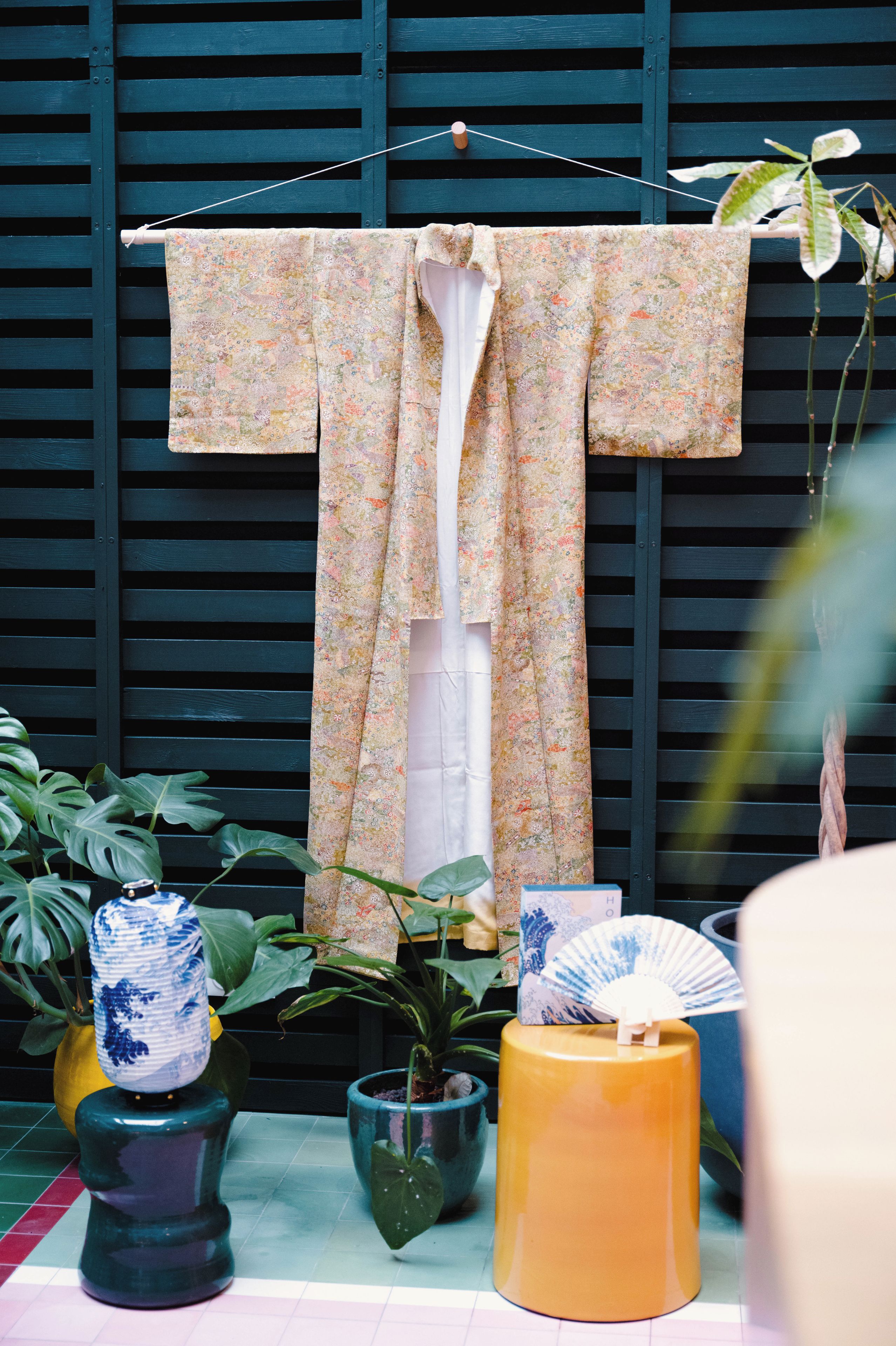 Una prenda tradicional japonesa colgada en una estructura de madera, rodeada de plantas en macetas y objetos decorativos.