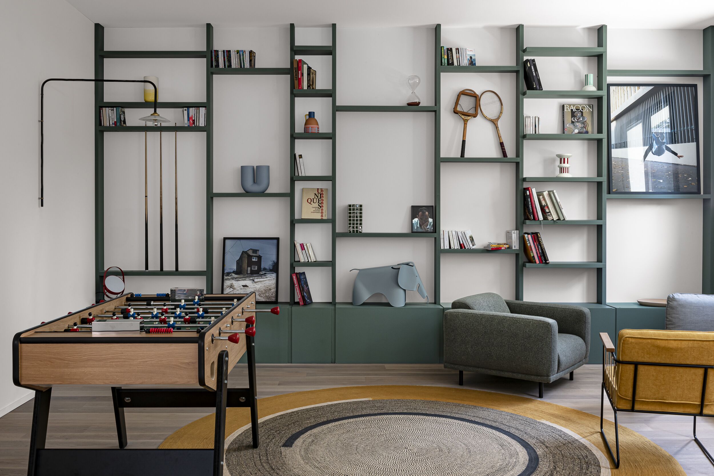 A modern living room with a foosball table, a grey sofa, and a bookshelf filled with books and decorative items.