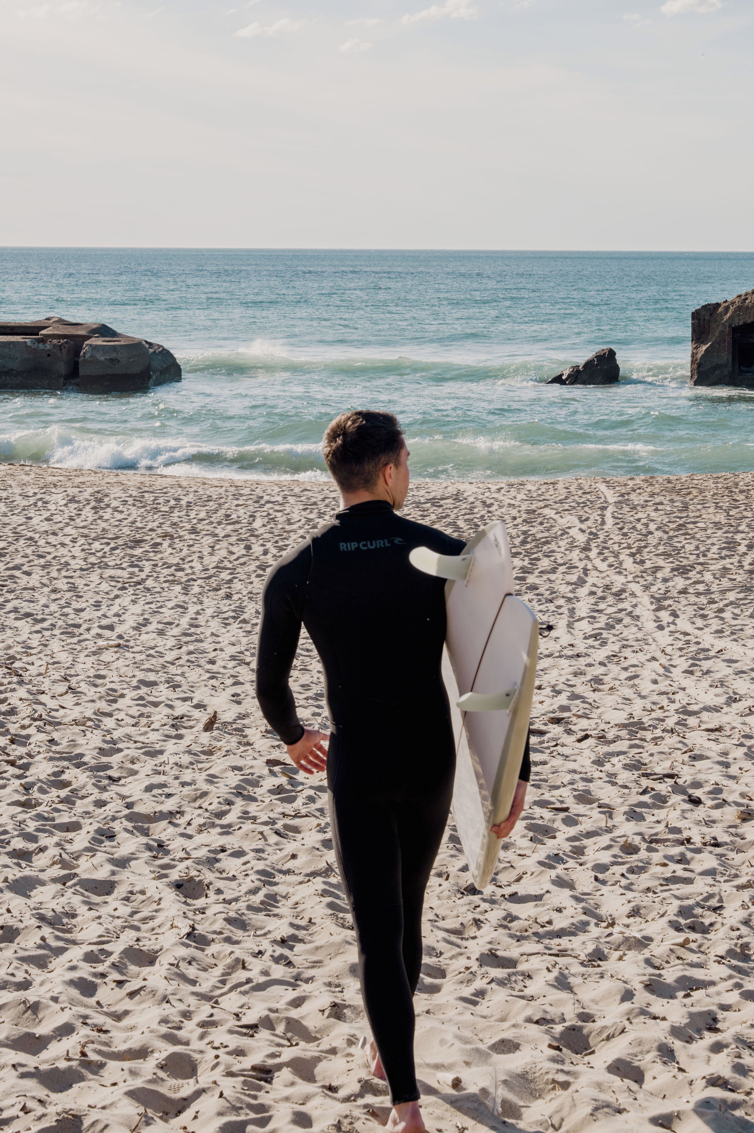 Una persona con traje de neopreno lleva una tabla de surf en una playa arenosa, con huellas visibles y olas al fondo.