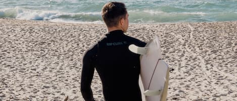 Una persona con traje de neopreno lleva una tabla de surf en una playa arenosa, con huellas visibles y olas al fondo.