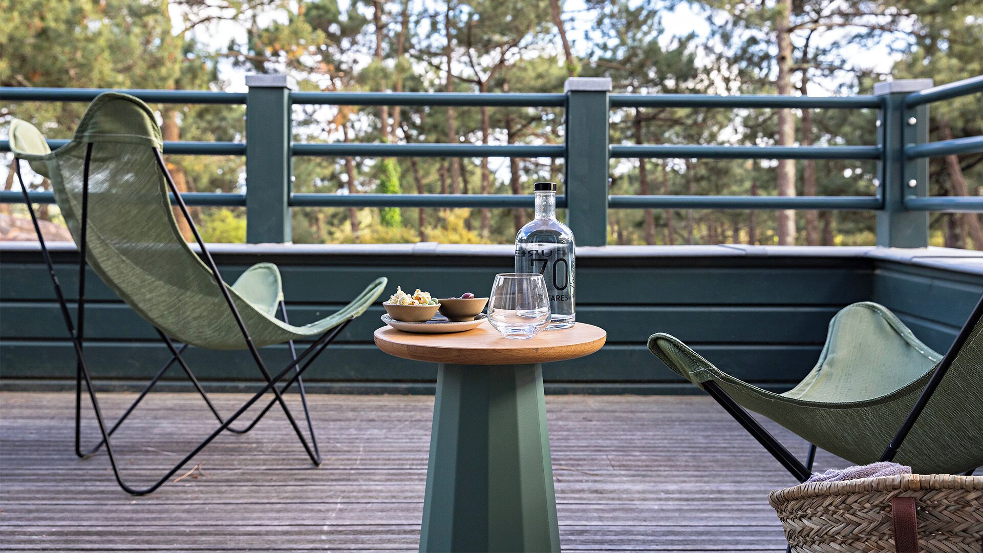 A wooden deck with a round table, two green chairs, a bottle, and a glass.