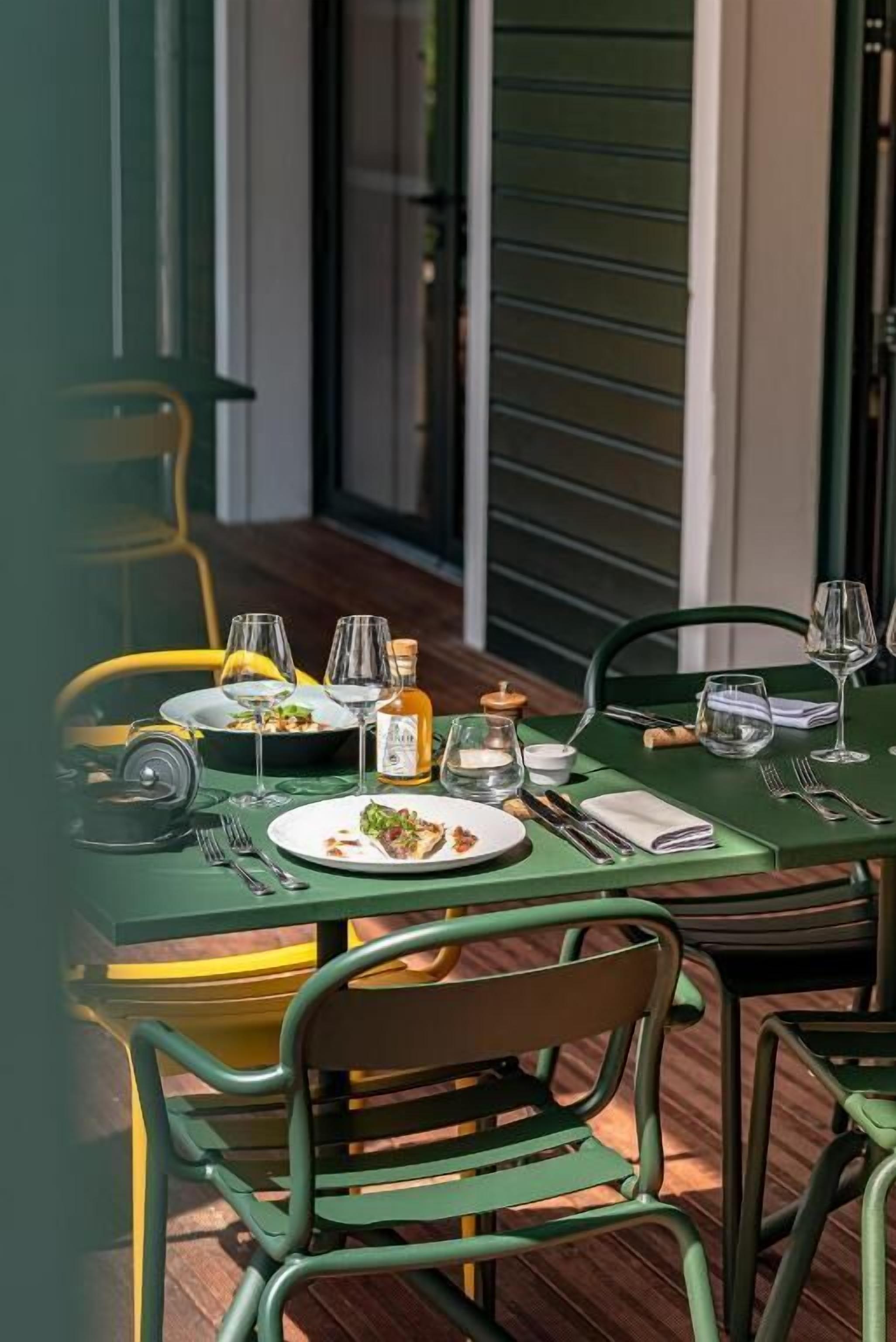 An outdoor dining area with green metal chairs and tables, set with plates of food, wine glasses, and cutlery.