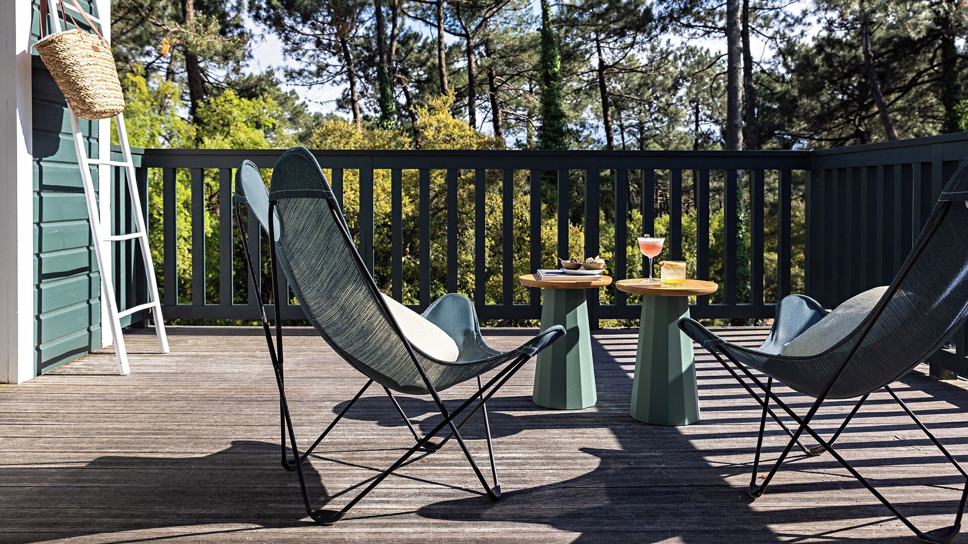 Eine Terrasse mit zwei Liegestühlen, einem kleinen Tisch mit Getränken und einem Korbwäscher auf einem Holzboden.