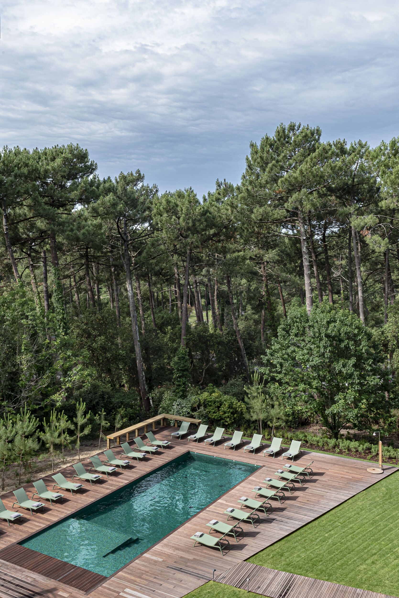 Piscina retangular com deck de madeira e espreguiçadeiras, cercada por uma área arborizada.