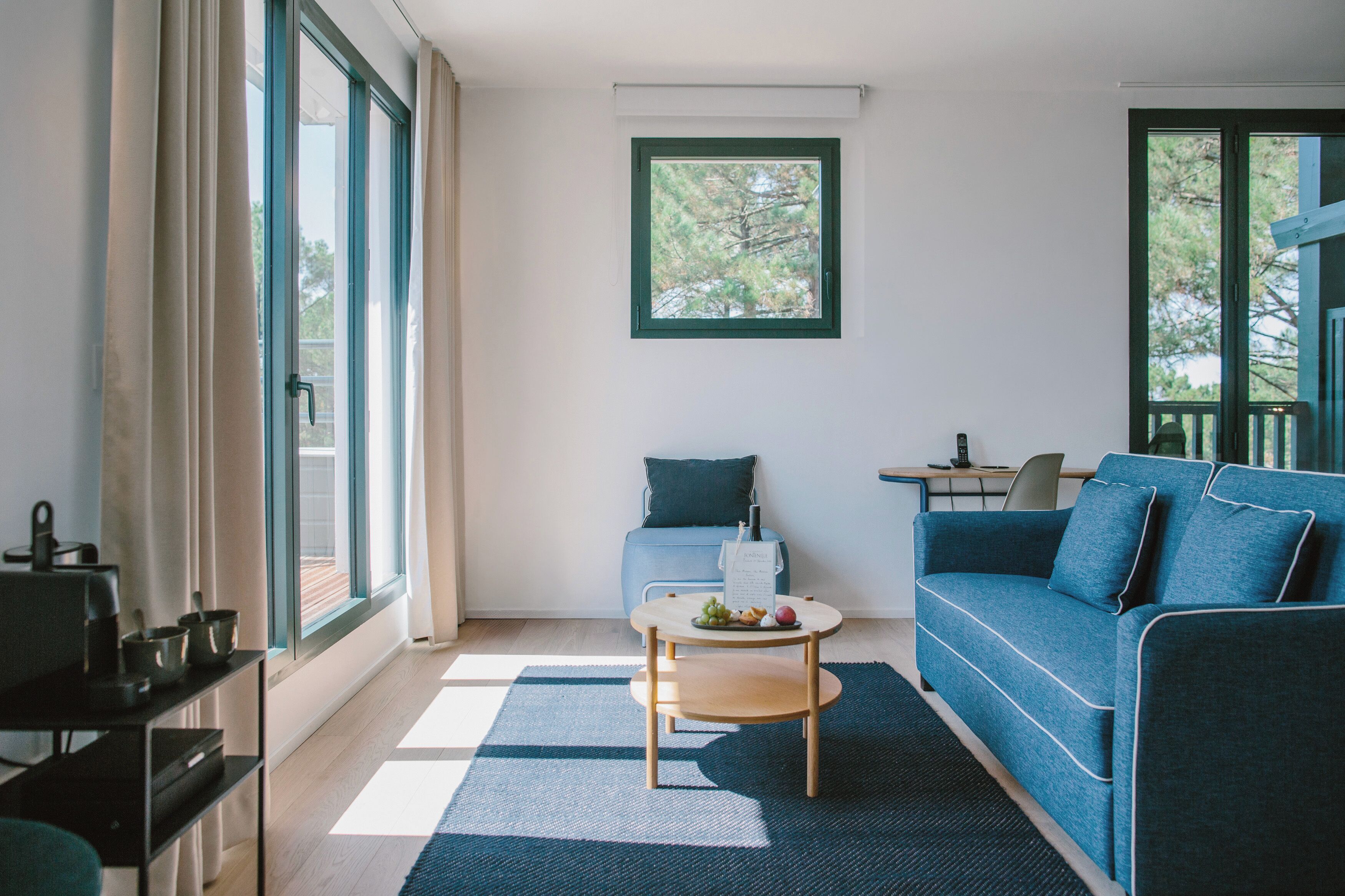 A modern living room with a blue sofa, a coffee table with fruits, and a view of trees through a window.