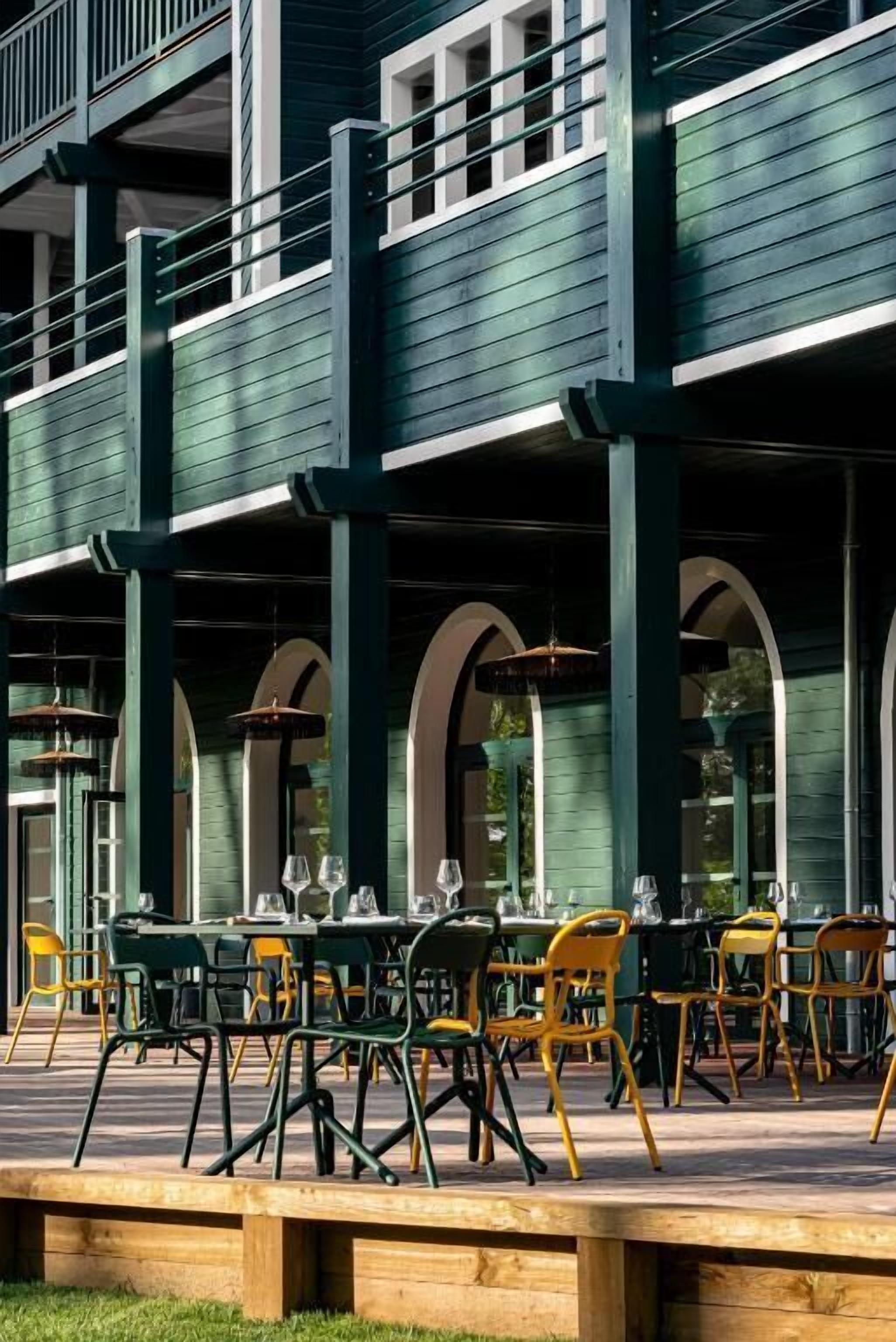 Un restaurant doté d’une terrasse, caractérisé par une architecture en bois vert et des chaises jaunes.