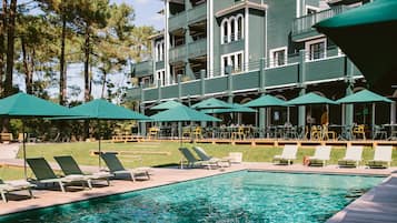 A pool area with lounge chairs and umbrellas, a building with balconies, and pine trees in the background.