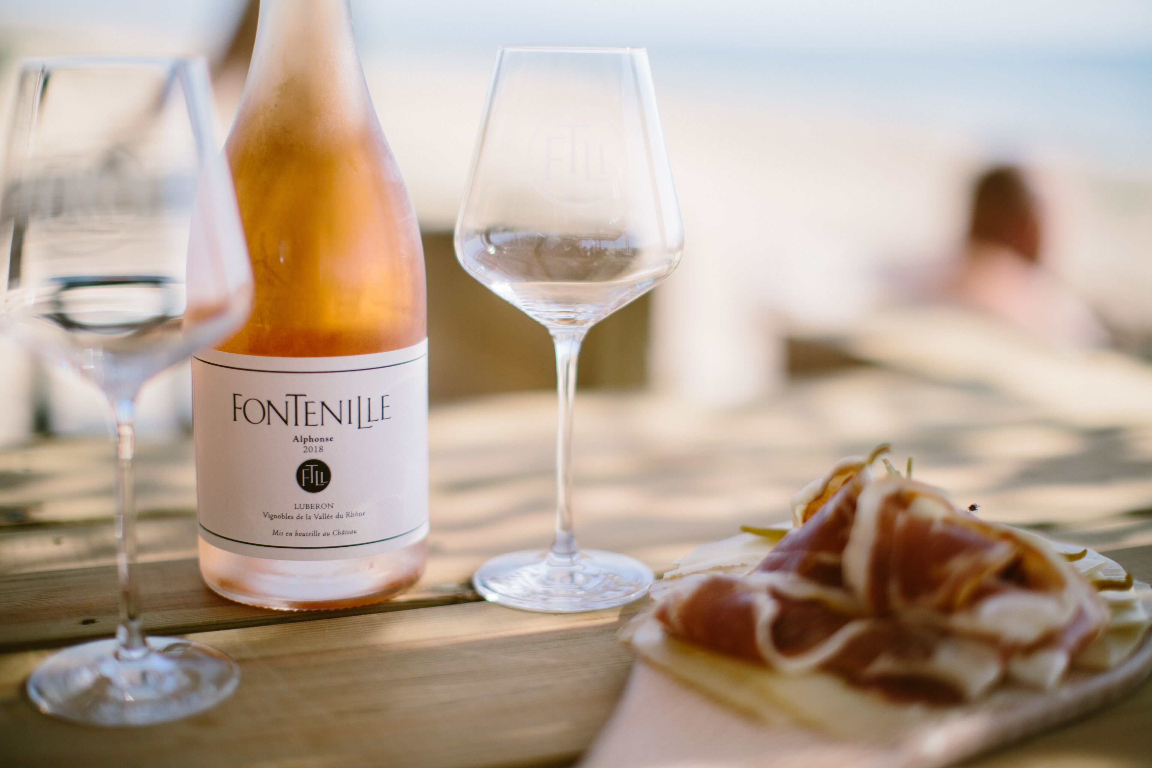 Una botella de vino Fontenille, dos copas y un plato de embutidos sobre una mesa de madera.
