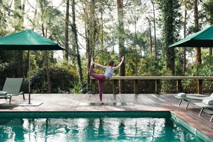 A person performing a yoga pose on a wooden deck by a pool with a forest backdrop.