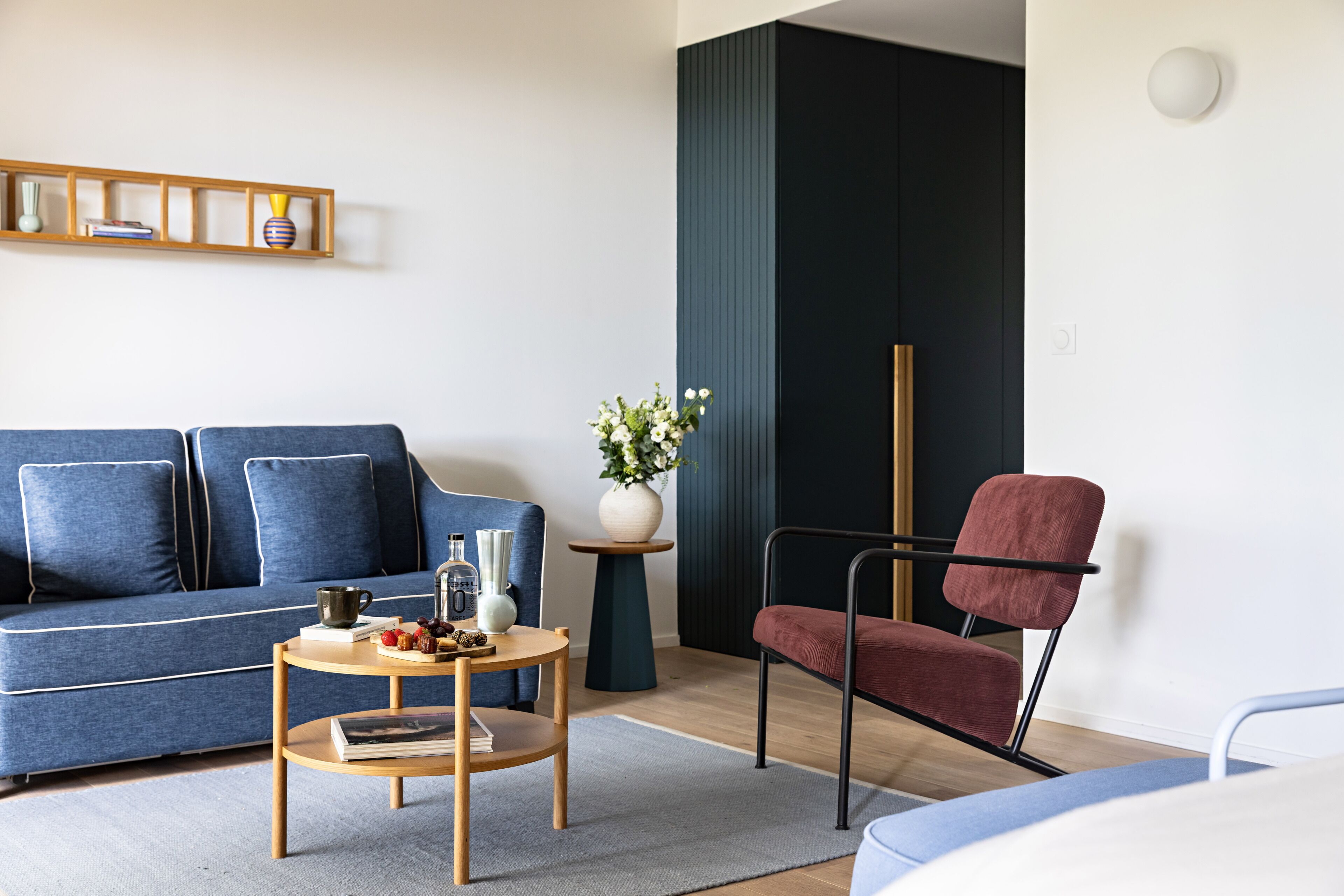 A modern living room with a blue sofa, a red chair, a small wooden table, and a vase with flowers.