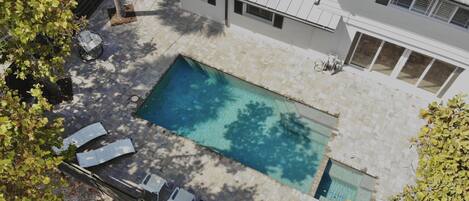 Una piscina rectangular con borde de baldosas, rodeada por un edificio blanco con techo de metal y ventanas.