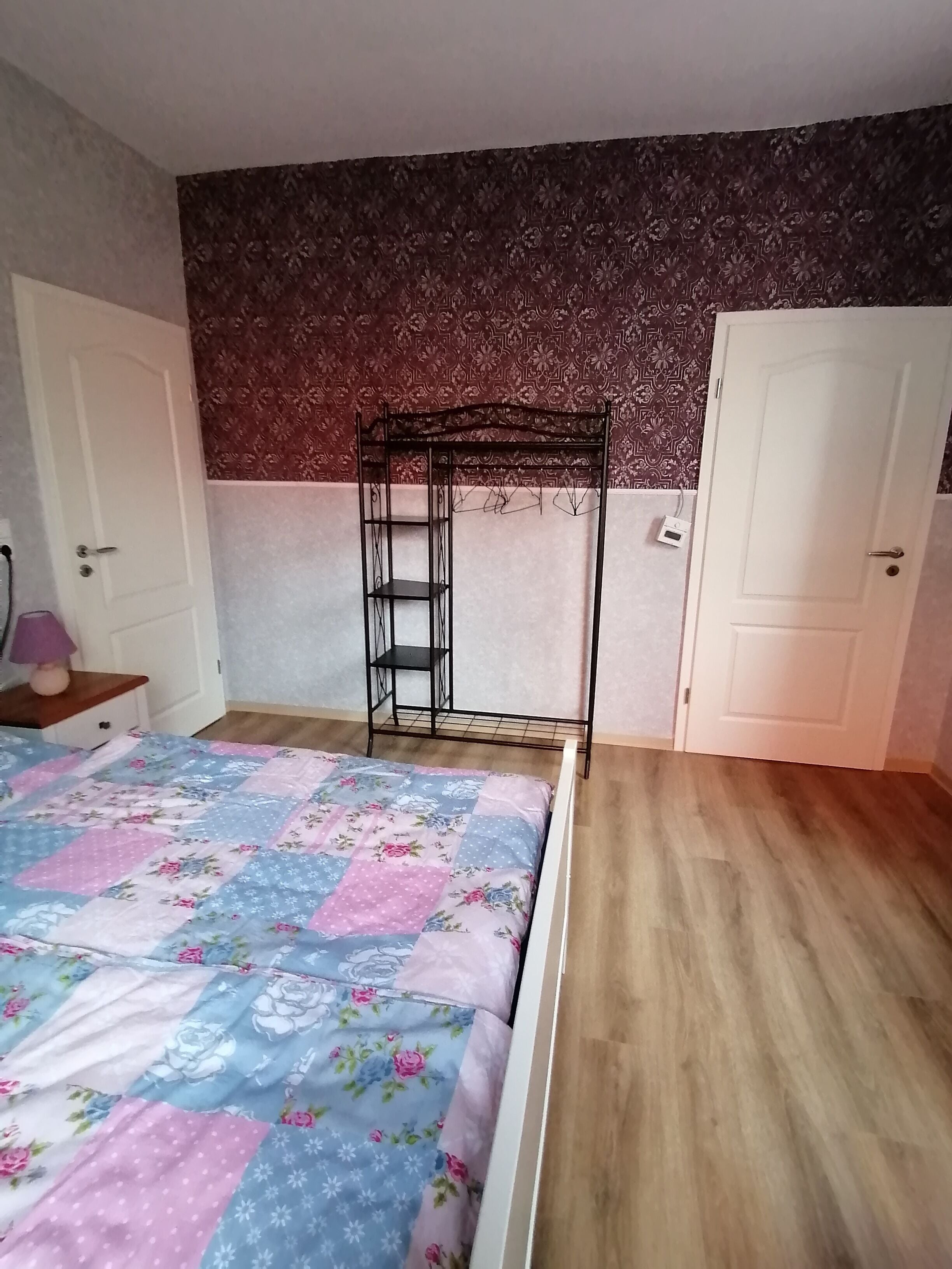 Un dormitorio con una cama, una estantería metálica, una puerta y papel tapiz estampado.