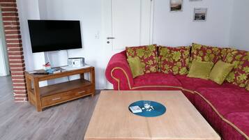 A living room with a wooden coffee table, a red sofa with floral cushions, a television on a wooden stand, and a door with a white frame.