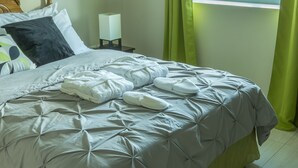 Een net opgemaakt bed met een grijze quilt en witte handdoeken, een nachtkastje met een lamp en groene gordijnen.