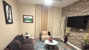 A modern living room with a sectional sofa, a round coffee table, a flat-screen TV mounted on a textured wall, and a ceiling fan.