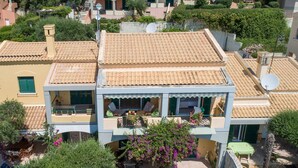 Ein zweigeschossiges Gebäude mit Balkon, einer Terrasse mit Gartenmöbeln und einem Gartenbereich mit blühenden Pflanzen.