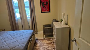 A hotel room with a bed, a desk, a chair, a radiator, and a window with curtains.