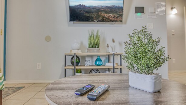 A living room with a round wooden table, two remote controls, a potted plant, and a flat-screen TV mounted on the wall displaying a landscape. - ✨ BY FORT BLISS: 2 KING BEDS, FAST WIFI, FULL BATH, LAUNDRY (El Paso)