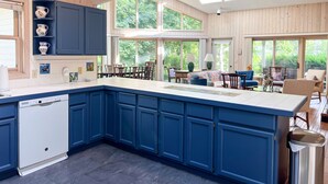 A spacious kitchen with blue cabinets, a dishwasher, and a large island. There are multiple windows providing natural light and views of the outdoors.
