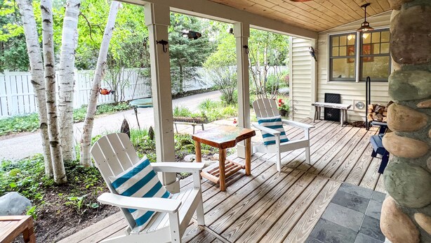 A covered porch with white Adirondack chairs, a small table, and a stone wall.