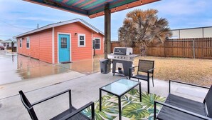 A covered patio with a grill, chairs, and a small house in the background. - Jewels Peace of Paradise (Rockport)