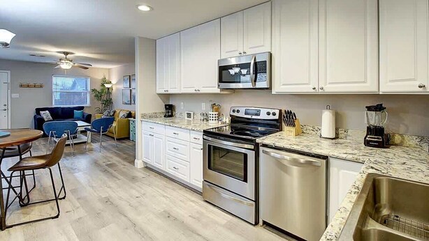 A modern kitchen with white cabinets, stainless steel appliances, a granite countertop, and a dining area with a wooden table and chairs. - Jewels Peace of Paradise (Rockport)