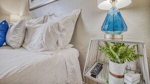 A neatly made bed with white bedding and a blue pillow, a bedside table with a blue lamp, a remote control, and a potted plant. - ☕ BY FORT BLISS: LAUNDRY, SECURITY SYSTEM, REFRIGERATION, KING BEDS (El Paso)