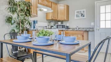 On aperçoit un coin repas avec une table en bois dressée pour quatre personnes, entourée de chaises. Au fond, on distingue une cuisine avec des placards en bois et un four à micro-ondes.