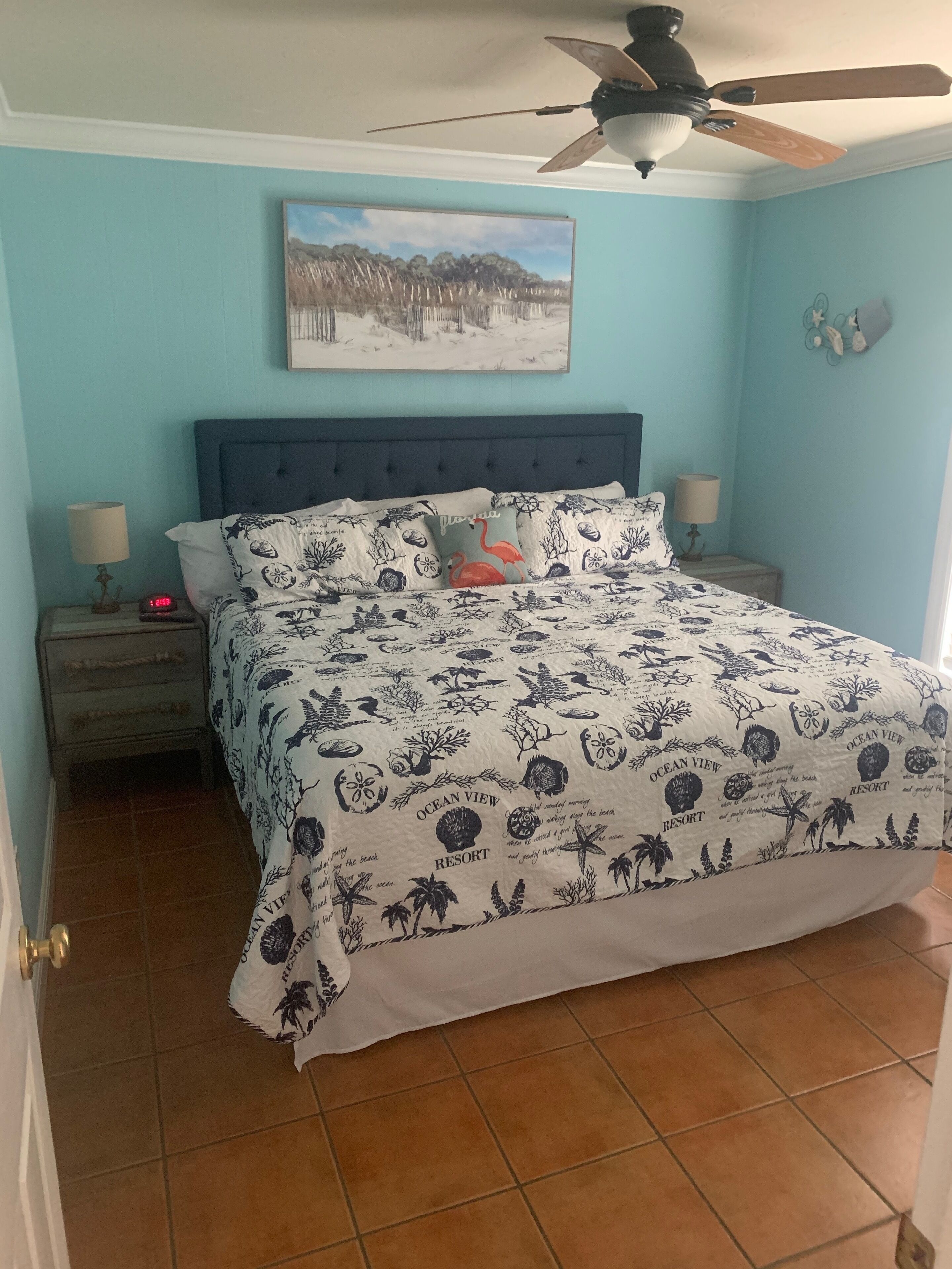 Une chambre à coucher comprenant un lit, une table de chevet, une horloge murale, un ventilateur de plafond et un tableau.
