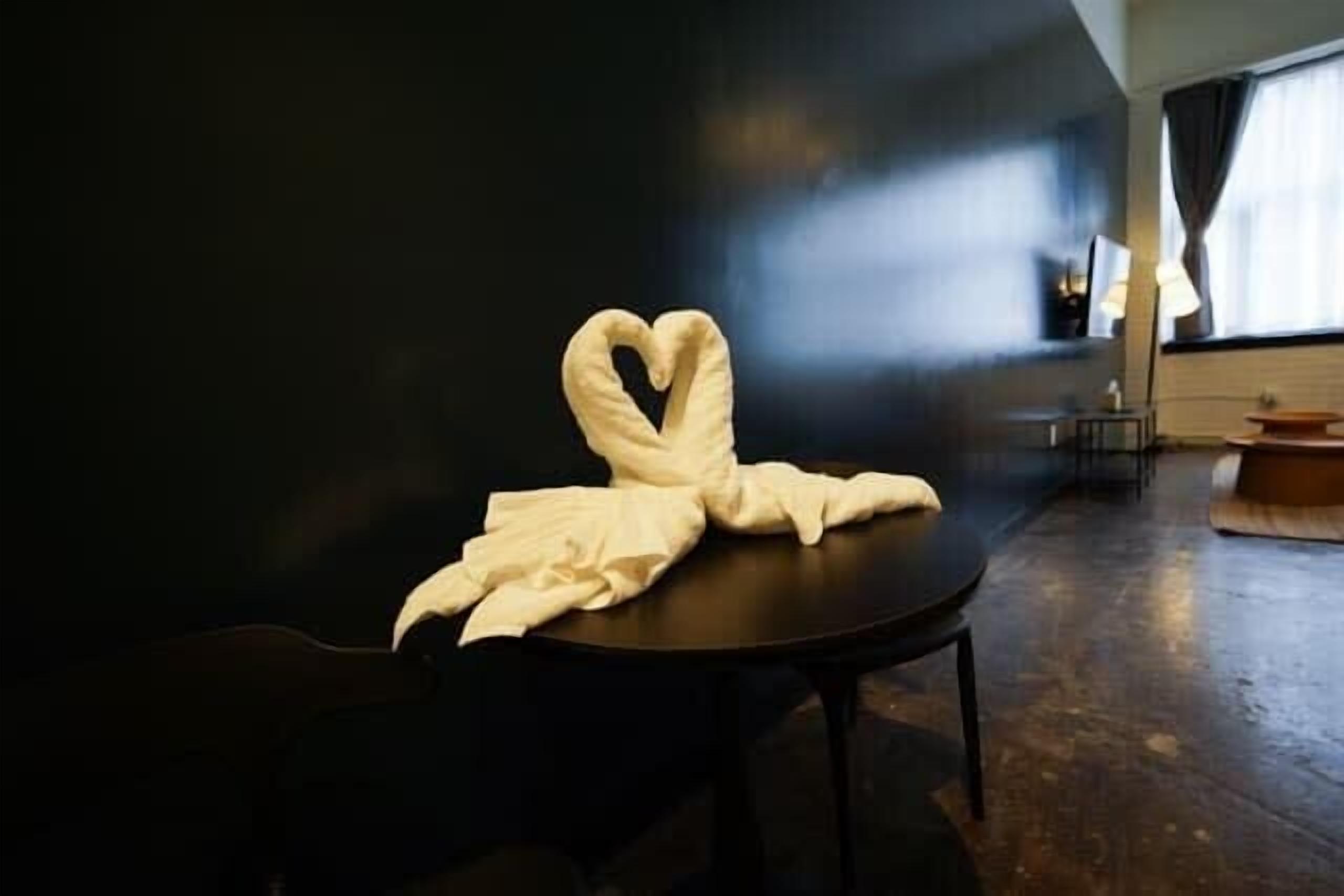 A room with a heart-shaped towel on a table, a lamp, and a window with curtains.