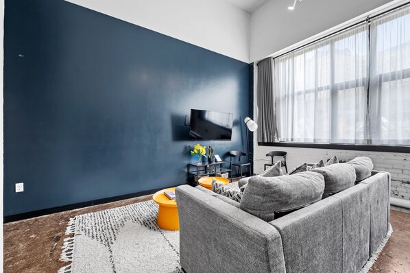 A modern living room with a dark blue accent wall, a grey sofa, a yellow coffee table, and a flat-screen TV mounted on the wall.