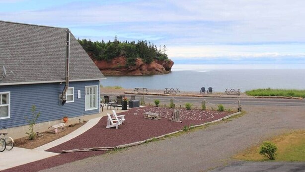 A building with a blue exterior, a paved driveway, outdoor seating, and a view of a body of water and a rocky shoreline.