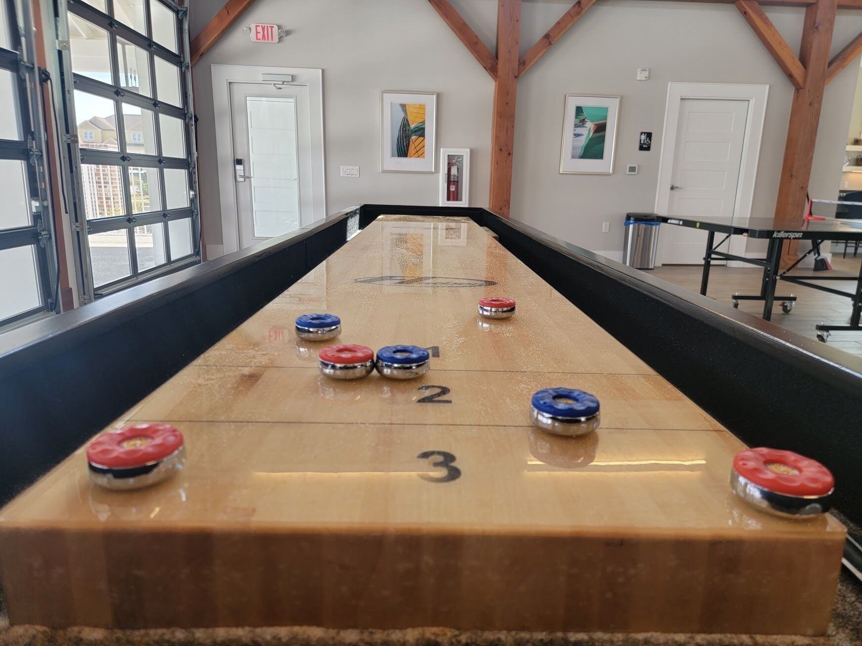 A shuffleboard table with numbered ends and pucks in a room with wooden beams, framed pictures, and exit signs.