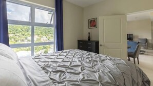 Een slaapkamer met een bed, een dressoir, een deur en een raam met uitzicht op groen.