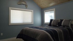 Une chambre avec un plafond en pente, deux fenêtres équipées de stores, un lit avec une literie à rayures et un élément décoratif mural.