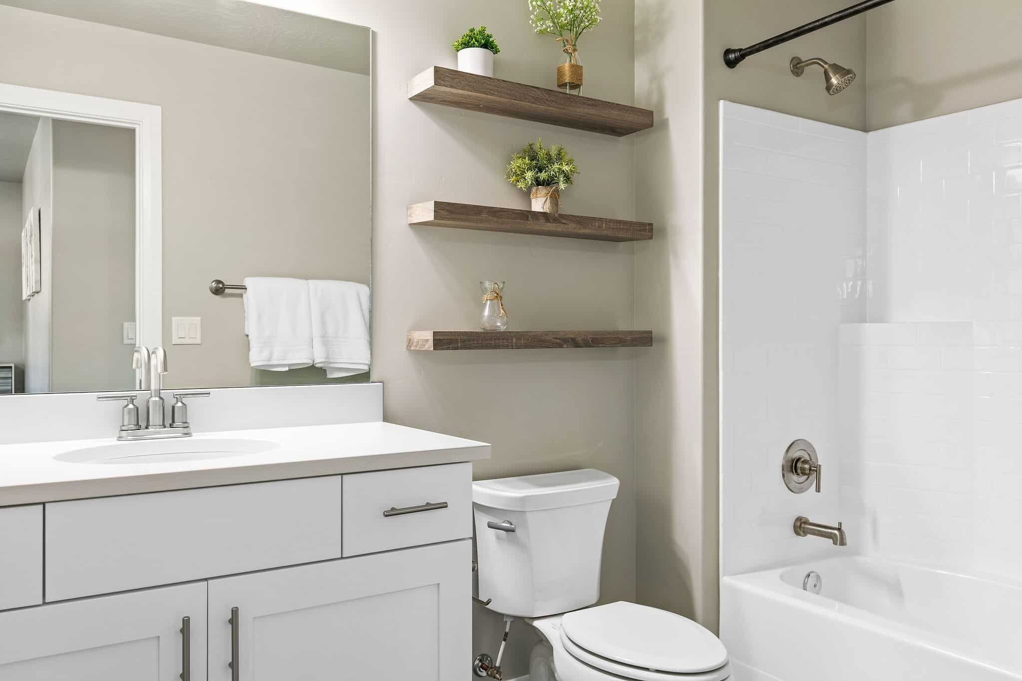 A modern bathroom with a white sink, toilet, and bathtub. There are wooden shelves with plants and a towel rack.