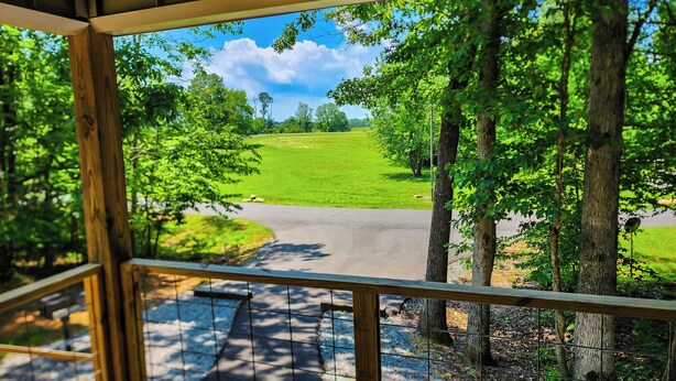 A view from a covered porch with a wooden railing, overlooking a green field and a road.
