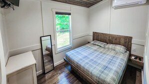 A bedroom with a wooden ceiling, a window with a view of greenery, a bed with a patterned bedspread, a bedside table, and an air conditioning unit.