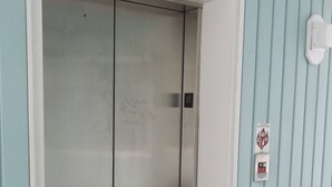 Ascensore con finitura metallica, montante bianco e pannello di controllo con simboli di emergenza e sicurezza antincendio.