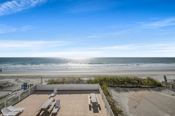 A beach view with a patio area featuring white lounge chairs and picnic tables. - Sea Aire II Duplex Up, Pet Friendly, Oceanfront, Shared Game Room (North Myrtle Beach)