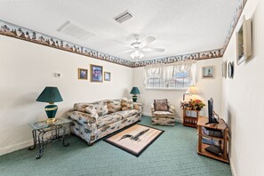 Ein Wohnzimmer mit einem gemusterten Sofa, einem kleinen Beistelltisch, einem Sessel, einer Stehlampe, einem Deckenventilator und einem Fenster mit Vorhängen.