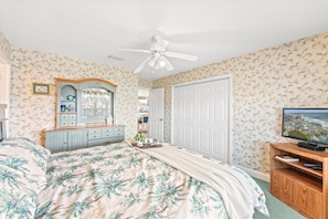 A bedroom with a bed, dresser, mirror, wardrobe, ceiling fan, and a television.